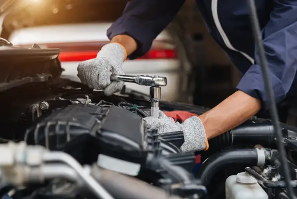 A mechanic wearing grey work gloves uses a ratchet wrench on an engine component under a car's open hood.