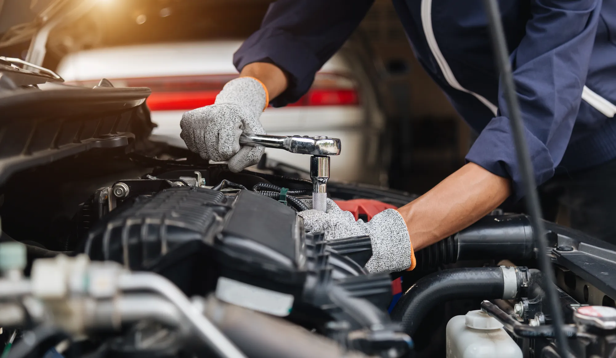 A mechanic wearing grey work gloves uses a ratchet wrench on an engine component under a car's open hood.