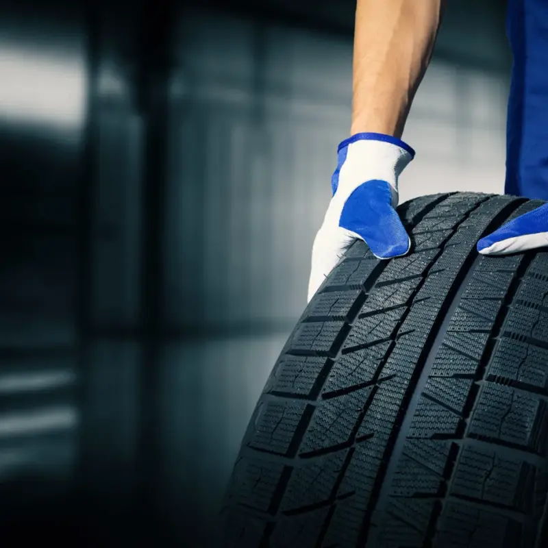 A mechanic in blue coveralls and work gloves rolls a new black tire across a garage floor.