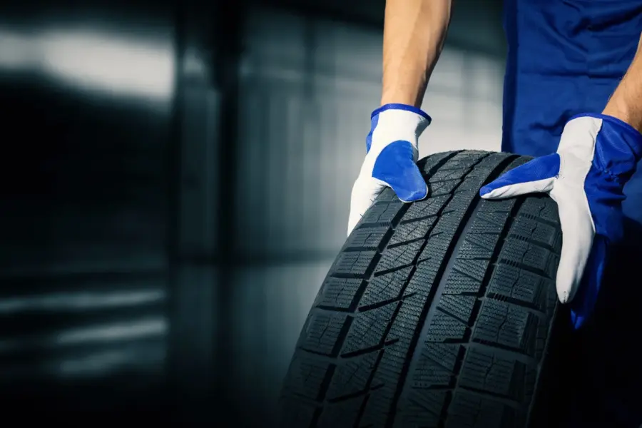 A mechanic in blue coveralls and work gloves rolls a new black tire across a garage floor.