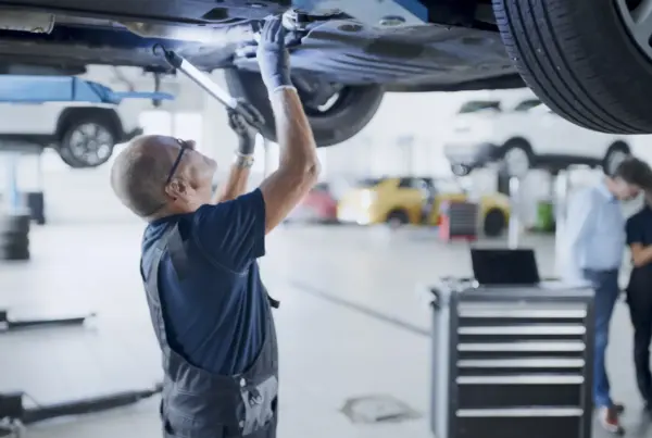 A mechanic in overalls and safety glasses works underneath a car raised on a lift in a busy auto repair shop.